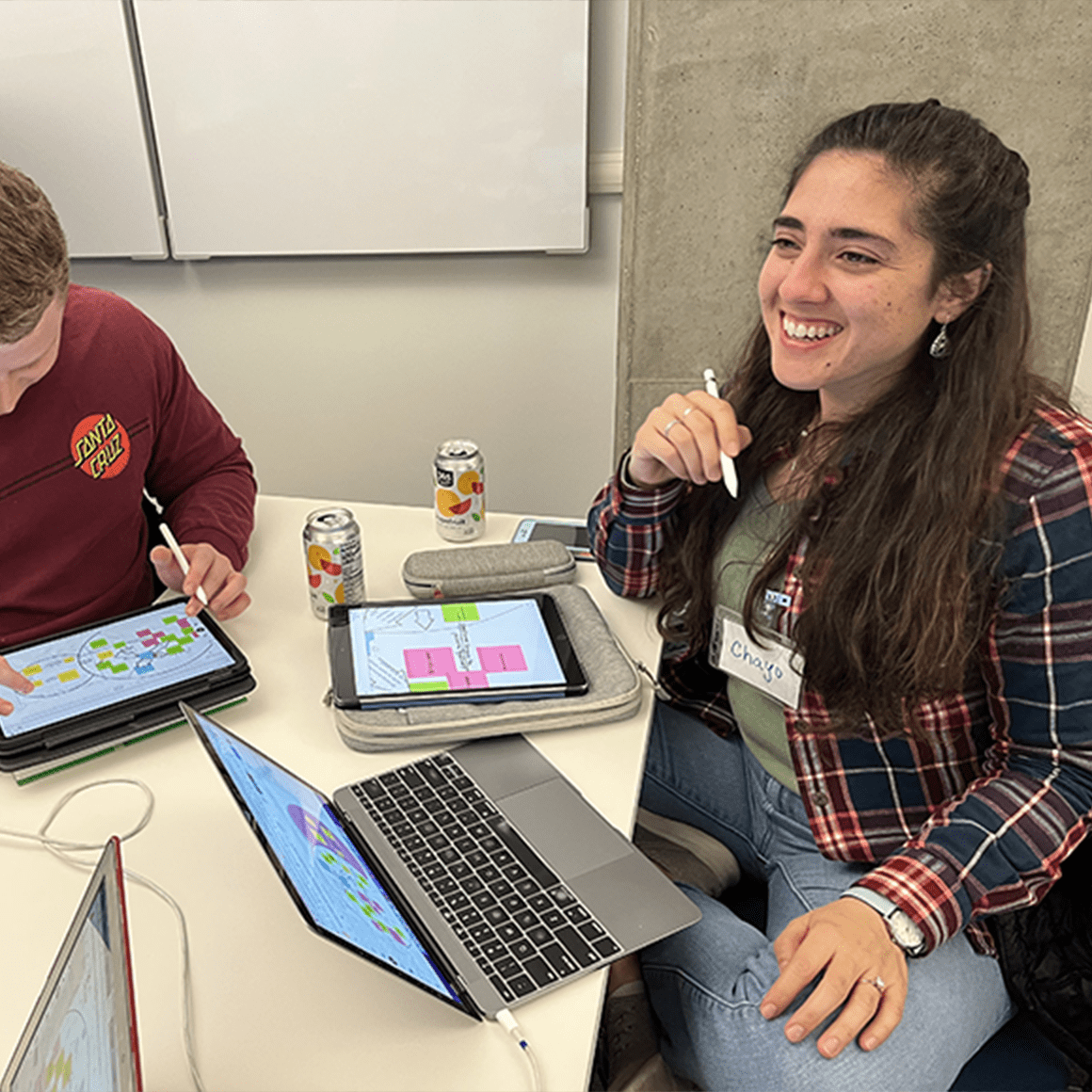 Two graduate students engaged in active learning. Students are smiling. There are computers and cans on the table.